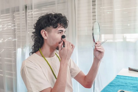 Young man with curly hair is applying makeup to his face with a brush. While holding a small mirror