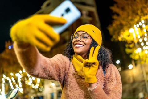 Smiling woman wearing yellow winter clothes taking selfie with smartphone and making peace sign in