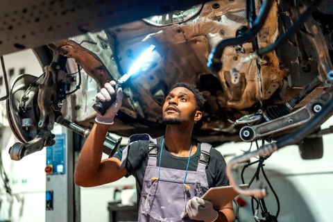 Young mechanic using flashlight and holding clipboard, inspecting car undercarriage in a