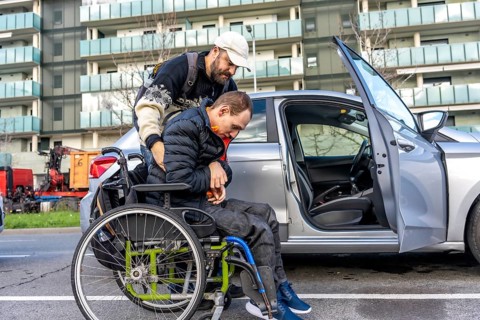 Caregiver assisting a disabled person getting into a car parked on a city street, showcasing