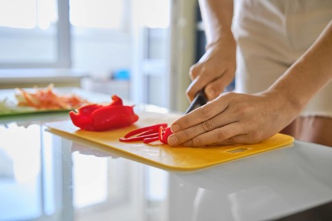 A person is slicing fresh red bell peppers on a yellow cutting board in a well-lit kitchen