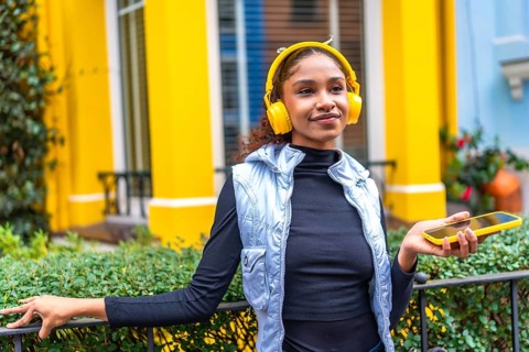Afro woman listening to music relaxes in the city leaning on garden rail of a colorful yellow house