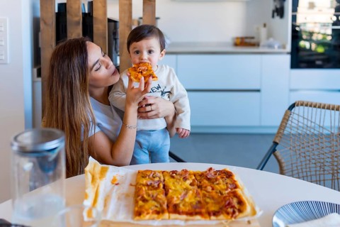 Mother enjoying a moment of togetherness while feeding her baby a slice of pizza at the kitchen
