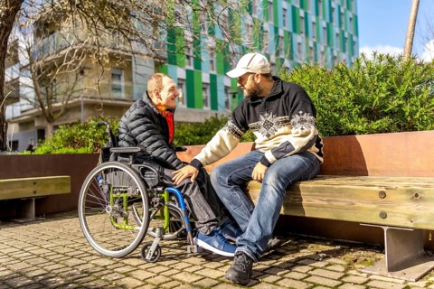 Two friends talking and smiling on a bench in a city park, one of them is using a wheelchair