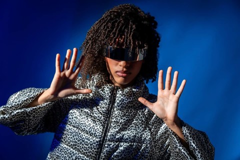 Stylish model wearing futuristic visor and patterned jacket pushing with hands on vibrant blue