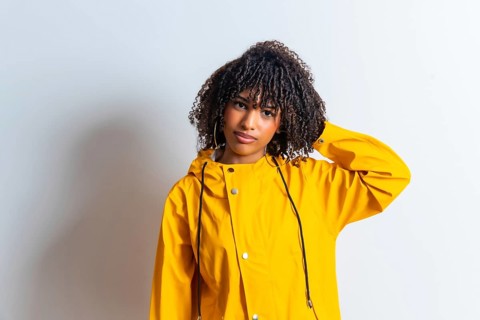 Studio portrait of a young woman wearing a yellow raincoat and scratching her head against a white