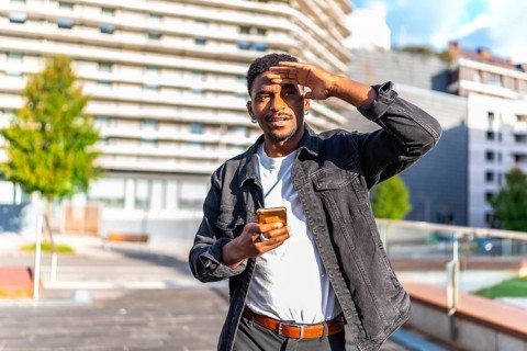 African man covering eyes from the sun while using mobile phone in the city searching for a