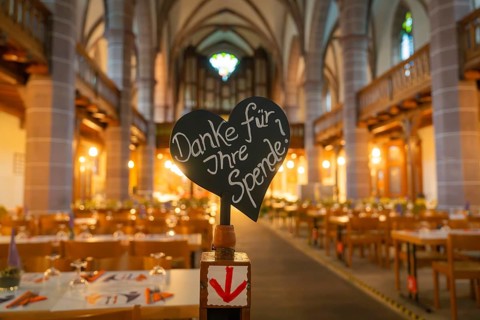 Appeal for donations in front of laid tables in a warmly lit church, Vesperkirche Calw, Black