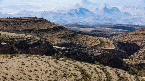 Slopes with barren vegetation and gorges, the mountains of the Hajar Mountains in the background,