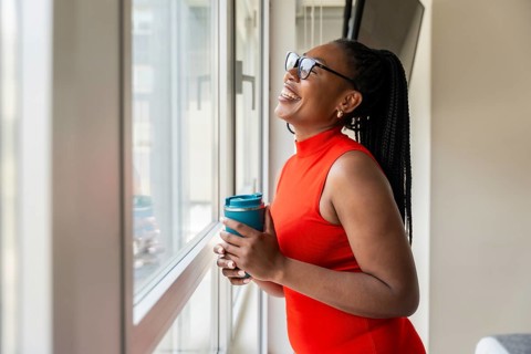 Happy businesswoman holding reusable coffee cup and looking out window during business trip,
