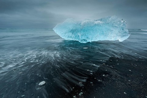 A shimmering blue iceberg is washed up by waves on a dark beach, Diamond Beach, Jökulsárlón glacier