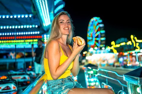 Blonde woman sitting eating a traditional hot dog at night fair
