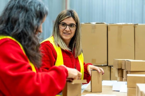 Two adult caucasian women talking relaxed while working in a distribution warehouse preparing boxes