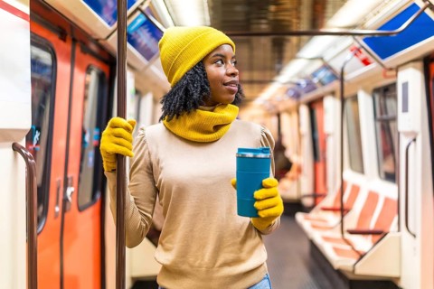 Young black woman wearing yellow winter clothes, holding a reusable coffee cup and traveling on a
