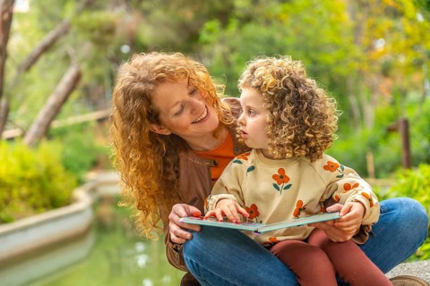 Cute girl and smiling caucasian mother reading a book sitting on a park together