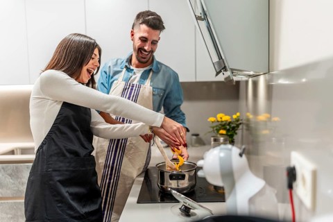 Happy couple wearing aprons preparing food in modern kitchen, stirring in pot and laughing