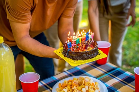Close-up of the hands of a young man holding birthday cake with candles during picnic with friends