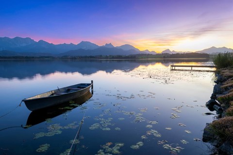 Boats at a jetty on a calm lake at sunset with mountains in the background, Hopfensee, Hopfen am