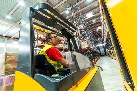 A worker in a yellow waistcoat is sitting driving a forklift in an industrial environment