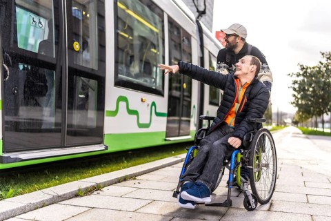 Young man in a wheelchair points at an approaching tram while his friend stands behind him,