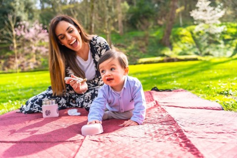 Mother giving food to her baby son during a picnic on a blanket in a park, surrounded by green