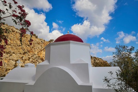 A white church with a red dome, surrounded by plants and rocks under a dramatic blue sky with