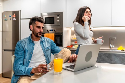 Young couple working from home in the kitchen, having breakfast and using laptop and smartphone