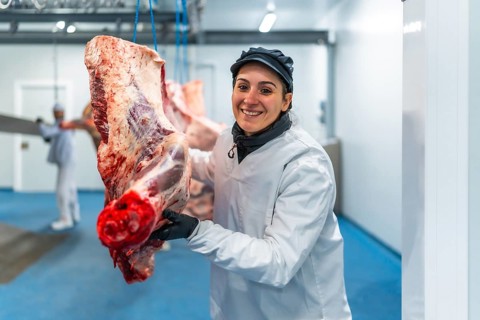 Happy female butcher carrying meat hanging in cold store of a slaughterhouse