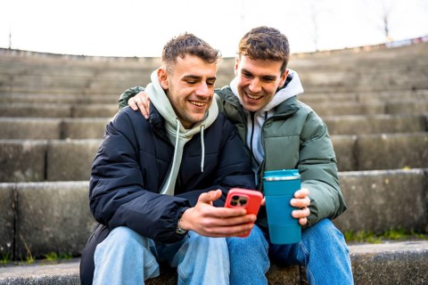 Two men are sitting on steps, embracing and looking at a smartphone while one holds a reusable