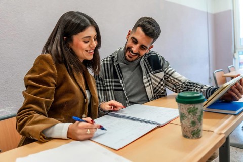 Two happy university students are studying together in a classroom, taking notes and using a