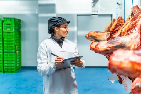 Woman in uniform and hat writing on clipboard checking goods in a meat processing factory