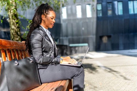 Latin beauty and elegant businesswoman working on remote using laptop sitting on a bench sunbathing