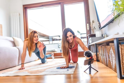 Two women in sportswear smiling and recording an online yoga class in their modern living room,