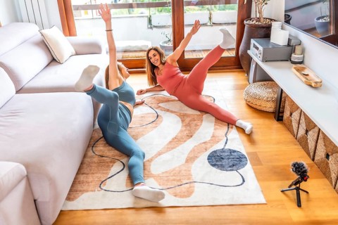 Two smiling women lying on carpet practicing yoga together at home following online class on