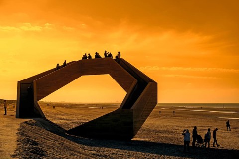 Westerpunt silhouetted against orange sunset sky, concrete sculpture on beach along North Sea coast