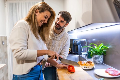 Cheerful couple making hamburgers together, slicing tomatoes and having fun in their modern kitchen