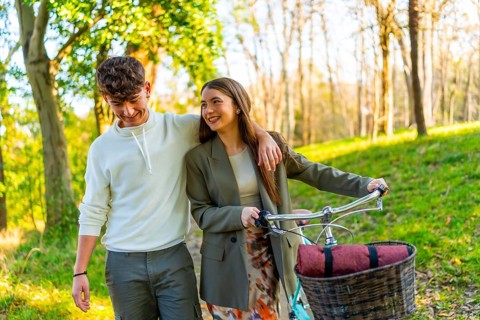 Smiling young couple walking in a sunlit park, sharing a moment together. Woman pushing a bicycle