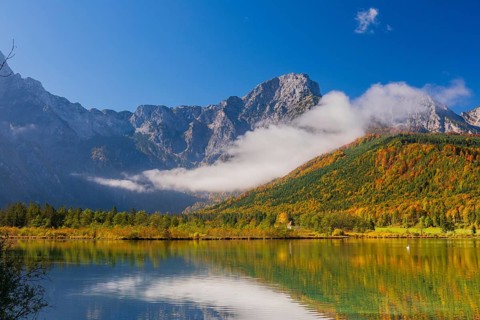 Lake Almsee in autumn with a forest reflected in the lake. A slowly dissipating fog moves over the