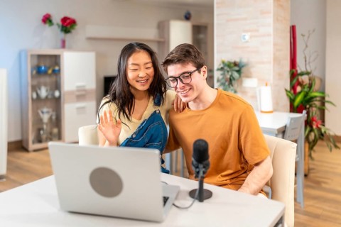 Happy multiethnic couple waving at the laptop camera while recording a vlog or streaming a podcast