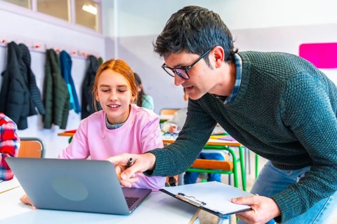 Teacher assisting female student using laptop during lesson in classroom, promoting interactive