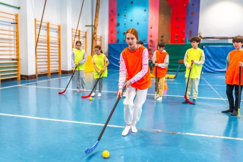 Group of elementary school students wearing orange and yellow vests playing floorball in a gym
