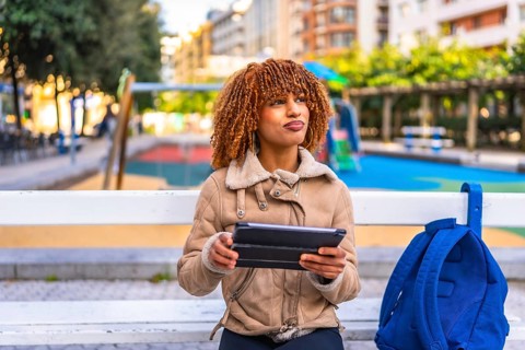 Pensive latin young woman in casual warm clothes using digital tablet sitting on bench in the city