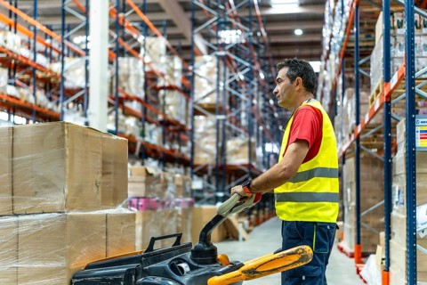 Side view of a mature man working in full warehouse moving boxes with low lift forklift