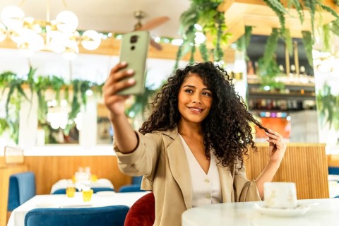 Sensual and happy latin woman taking selfie while touching her long curly hair sitting alone in a