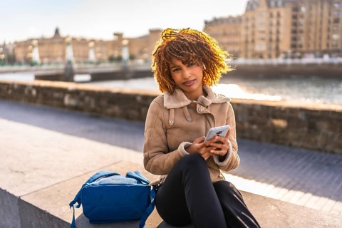 Beauty latin female student using smartphone sitting next to rucksack on the city during sunset