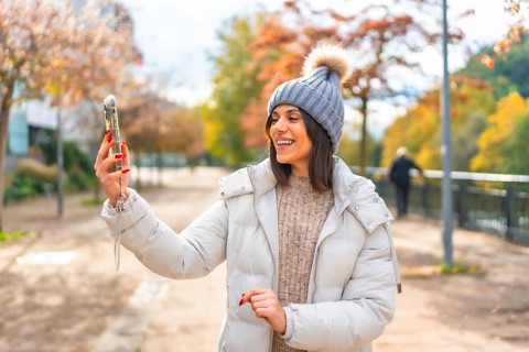 Woman wearing warm clothes and woolly hat smiling while talking during video call with mobile phone
