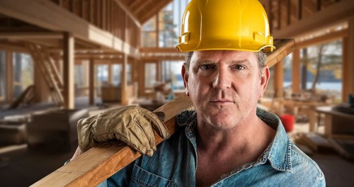 Male contractor at a construction site wearing a hard hat and work gloves holding lumber 2x4