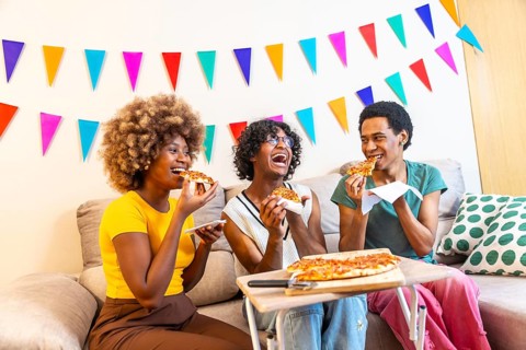 Diverse friends eating a slice of pizza sitting on the sofa celebrating at home