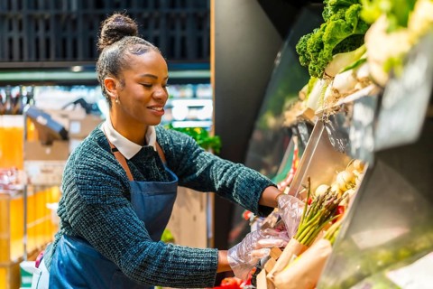 Smiling saleswoman wearing gloves arranging fresh organic asparagus on display in a supermarket