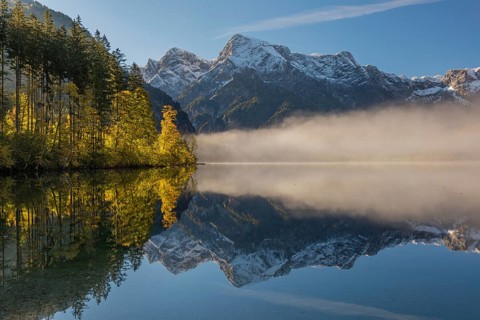 Lake Almsee in autumn with a forest and mountains reflected in the lake. A slowly dissipating fog